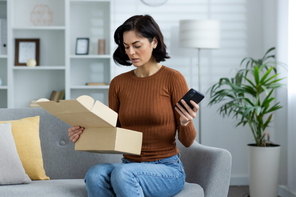 woman is sitting on the sofa at home, holding a mobile phone in her hand and worriedly looking into an open box of a parcel